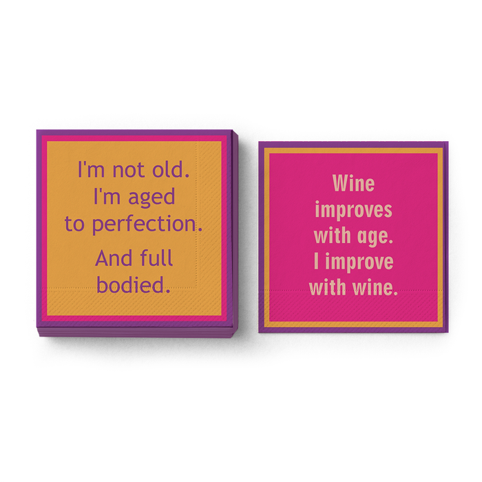 The Drinks On Me "Wine Improves With Age Napkin" set includes two colorful square cocktail napkins—one gold with purple "I’m not old. I’m aged to perfection." text, and one pink with white "Wine improves with age. I improve with wine.