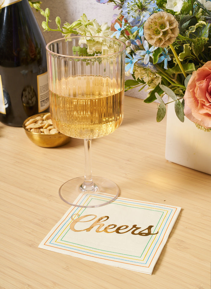 A Sophistiplate LLC Wine Modern Stemmed glass (17 oz, clear) from the 4-pack sits on a wooden surface beside a "Cheers" napkin, with sparkling wine inside. Nearby are a bowl of nuts, champagne bottle, and assorted flowers.