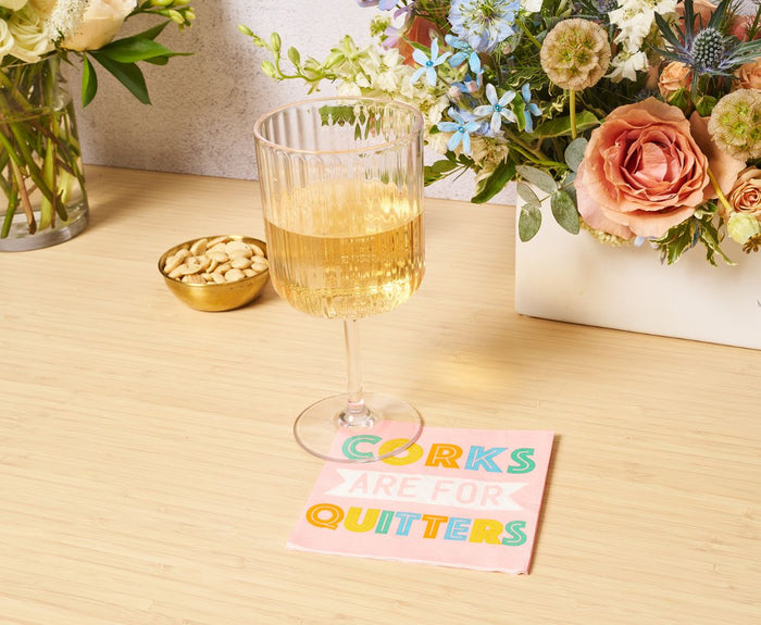 A Sophistiplate Wine Modern Stemmed Clear glass (17 oz) from the 4 Pack sits on a wooden table beside a bowl of nuts and a pink "CORKS ARE FOR QUITTERS" napkin, with colorful flowers in the background.