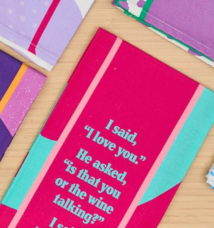 A pink notebook displays the witty “Is that you or the wine talking?” quote from the Drinks On Me Wine Talking Dish Towel, lying among colorful notebooks on a light wooden surface.