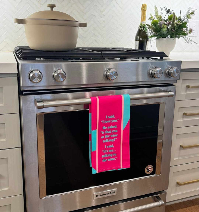A stainless steel oven shows the "Wine Talking Dish Towel" by Drinks On Me, a humorous kitchen gift. A beige pot sits on the stovetop, while flowers and a bottle of wine decorate the nearby countertop.