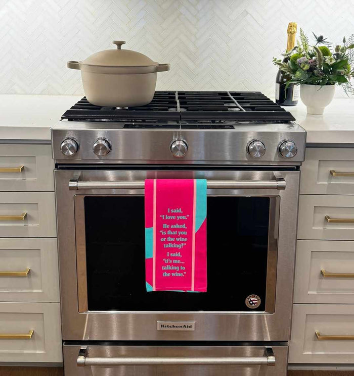 A beige Dutch oven sits on a stainless steel stove. The Drinks On Me “Wine Talking” dish towel, featuring a bright pink and teal design and humorous quote, hangs on the oven door. White cabinets and a floral arrangement are in the background.