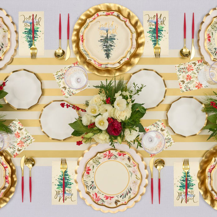 A festive table is set with Sophistiplate Winter Blossom Paper Salad Plates (8pk), red and gold cutlery, floral napkins, a centerpiece of white and red flowers, and holiday decor on a gold and white striped tablecloth.