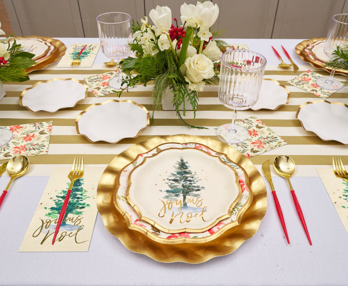 The Winter Blossom Table Setting by Sophistiplate creates a festive Christmas table with scalloped plates, Joyeux Noel napkins featuring a Christmas tree, gold cutlery with red handles, and a white floral centerpiece.