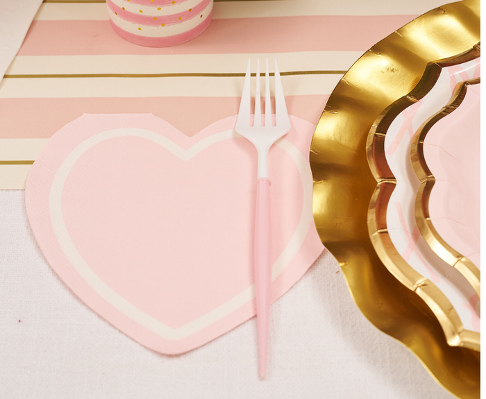 A pink heart-shaped Sugarplum XOXO Die Cut Lunch Napkin sits under a pink and white fork, next to gold-edged scalloped plates on a table with a pink and beige striped runner.