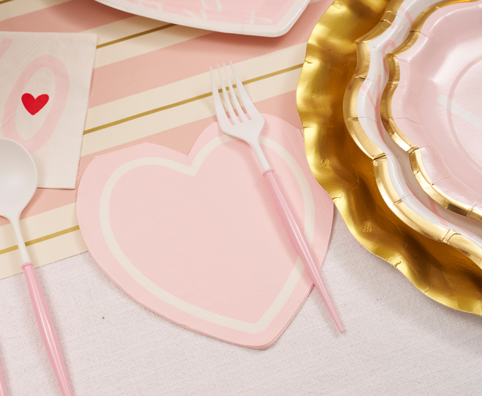 A pink and gold table setting with a heart plate, pink-handled utensils, XOXO collection scalloped plates, and Sugarplum XOXO Die Cut Lunch Napkins decorated with red hearts on a striped tablecloth.