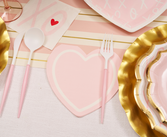 A pink and gold table setting with heart-shaped plates, pink utensils, a Sugarplum XOXO Die Cut Lunch Napkin, and a pink-striped runner creates a romantic and festive atmosphere.