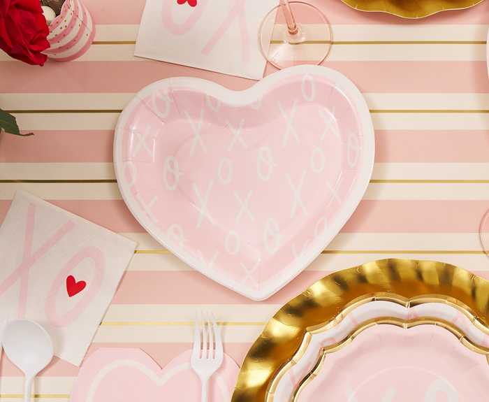 A Sugarplum XOXO Die Cut Salad Plate from the 8pk, heart-shaped and pink with white XO patterns, is set on a striped cloth among heart napkins, gold-edged plates, plastic cutlery, and a wine glass for a romantic Valentine’s Day.