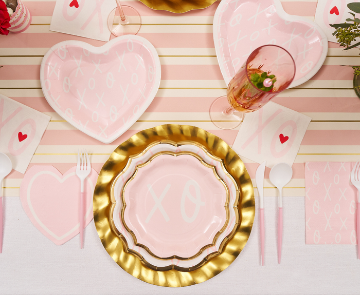 A festive table set with pink heart-shaped plates, Sugarplum XOXO Salad Plates (8pk), a gold-edged charger, pink utensils, heart napkins, a champagne glass with a strawberry, on a pink and white striped runner.