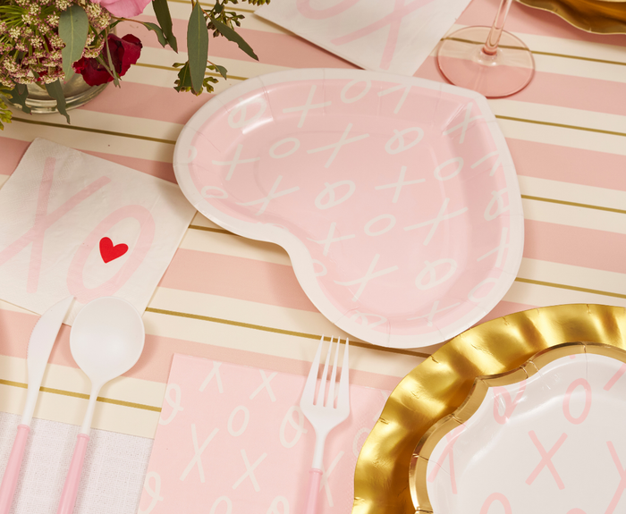 A Sugarplum XOXO Die Cut Salad Plate, heart-shaped and pink with white XO patterns, sits on a striped cloth amid matching Valentine’s plates, pink-and-white utensils, an XO napkin with a red heart, flowers, and a wine glass.