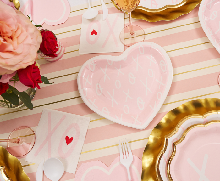 A pink and white striped table features Sugarplum XOXO Die Cut Salad Plates, gold-rimmed disposable party plates, plastic cutlery, XO napkins with a red heart, and a bouquet of red and pink roses.