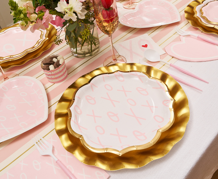 A festive table is set with Sugarplum XOXO Dinner Plates (8pk), gold-trimmed disposables, pink utensils, flowers, and a decorated drink. XO patterns on the runner create a romantic, playful Valentine’s Day atmosphere.
