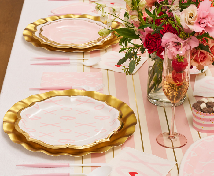 A festive table setting with pink and gold plates, floral napkins, Sophistiplate Blush Bella Plastic Cutlery (24pc), a vase of pink and red flowers, champagne with raspberries, heart decor, and a pink-striped table runner.