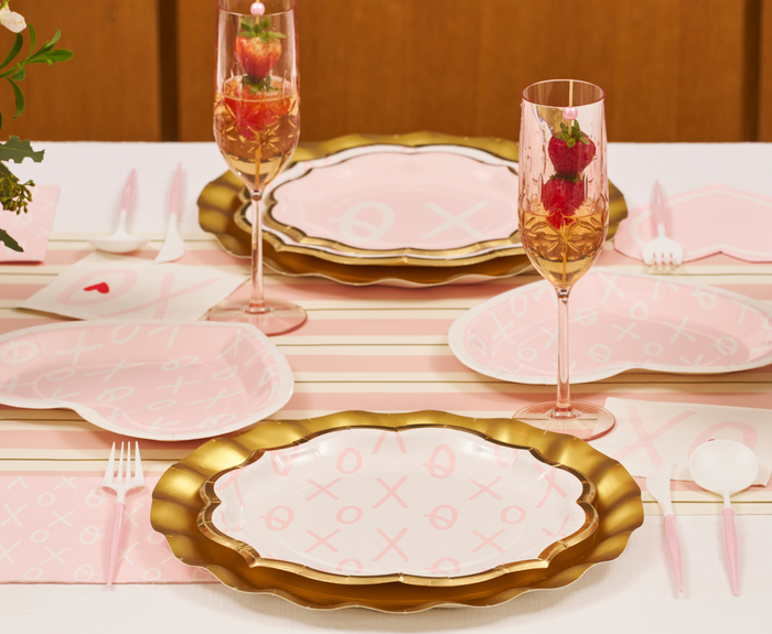A table set with Sugarplum XOXO Dinner Plates (8pk), gold-rimmed pink disposable plates, heart-themed napkins, pink strawberry glasses—all arranged on a striped table runner for Valentine’s Day.