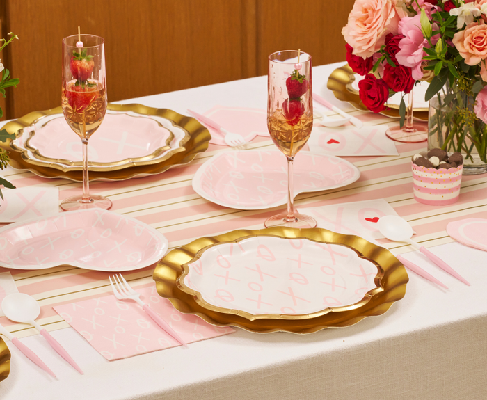 A festive table set with Sugarplum XOXO Dinner Plates (8pk), pink cutlery, champagne glasses garnished with strawberries, and pink and red rose arrangements creates a romantic Valentine’s Day theme.