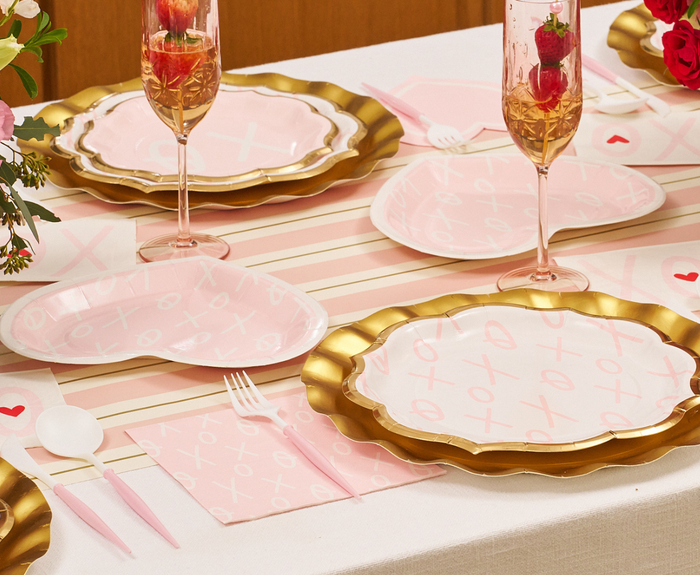 A table set for a celebration with Sugarplum XOXO Dinner Plates (8pk), gold charger plates, pink utensils, champagne flutes garnished with strawberries, and a striped tablecloth.