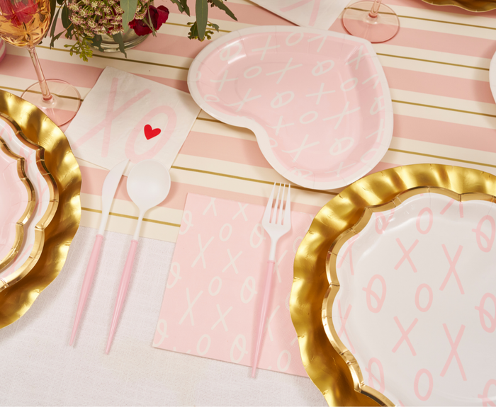 A festive table features a pink and white striped tablecloth, gold-edged heart and round plates with XO motifs, pink cutlery, Sugarplum XOXO Lunch Napkins/20pk, and wine glasses filled with pink drinks.
