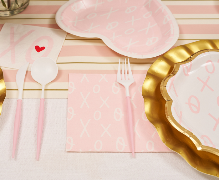 A romantic Valentine’s table features pink and gold plates, pink cutlery, a striped runner, and Sugarplum XOXO Lunch Napkins (20pk). Accents include a white napkin with a red heart for a soft, love-inspired theme.