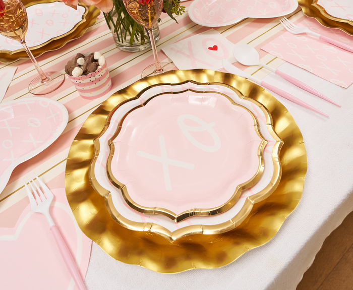 A pink and gold scalloped plate set is displayed on a table with Sophistiplate Blush Bella Plastic Cutlery (24pc, Service for 8), pink napkins, heart-shaped placemats, XO decor, and a small cup of chocolates.