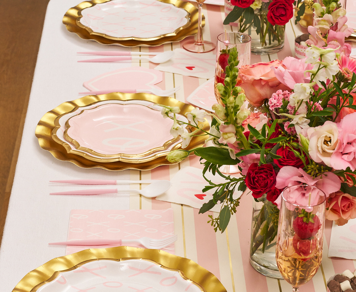A table set with Sugarplum XOXO Salad Plates, pink napkins, pink cutlery, drinks in glasses, and bouquets of pink and red flowers as centerpieces on a white tablecloth with gold stripes.