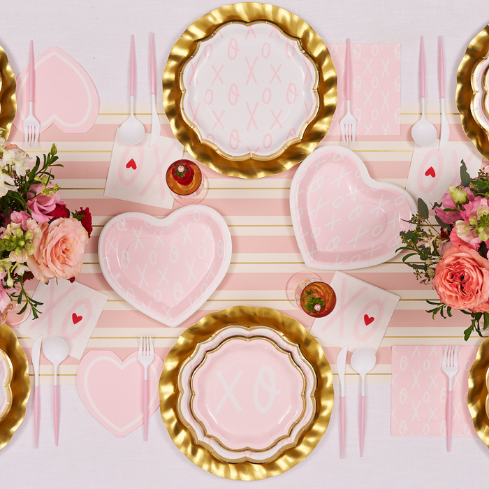 Create a romantic Valentine’s Day table with Sugarplum XOXO Dinner Plates (8pk), gold accents, heart-shaped dishes, striped placemats, pink utensils, floral centerpieces, and napkins decorated with red hearts.