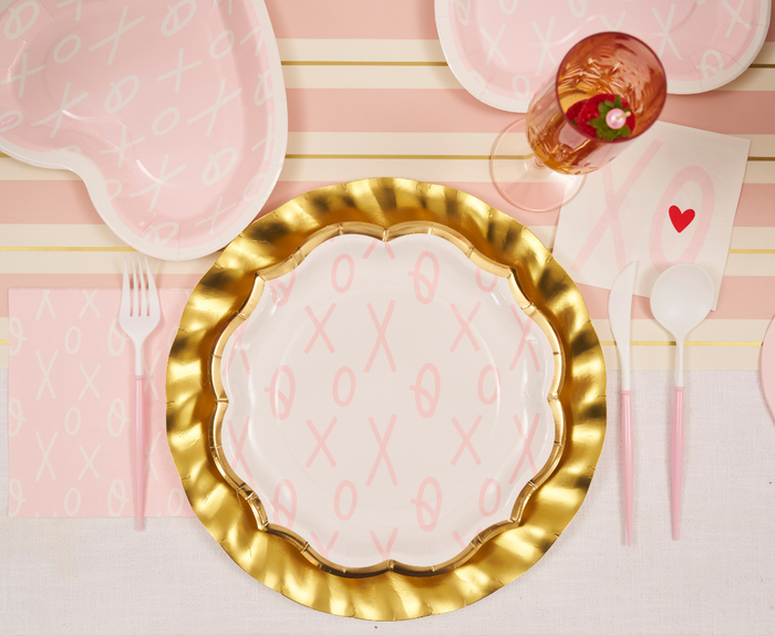 A table is set with Sugarplum’s XOXO Dinner Plates (8pk), featuring pink X and O patterns, paired with a gold scalloped charger, pink cutlery, a pink napkin, and a pink drink for a festive Valentine’s look in pink, white, and gold.