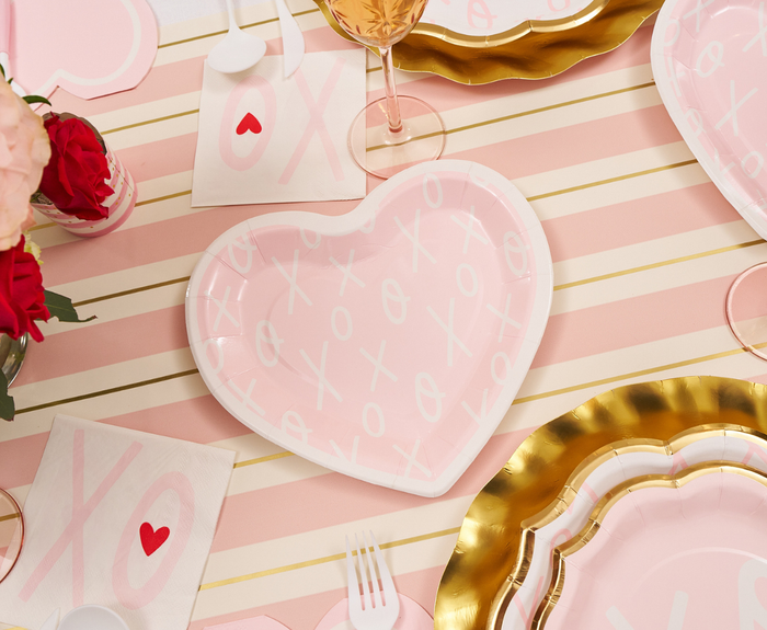 A heart-shaped pink plate with XO patterns rests on a pink and white striped tablecloth with gold lines. Gold-rimmed plates, Sophistiplate XOXO Cocktail Napkins (20pk), plastic utensils, and a wine glass complete the festive setting.