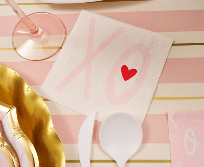 The Sophistiplate XOXO Cocktail Napkins/20pk feature large pink XO letters and a red heart—stylish white napkins ideal for your Valentine’s Day gathering, shown on a pink and white striped table with gold accents.