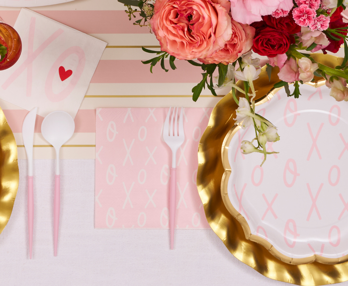 A festive table is set with gold-rimmed plates featuring pink XO patterns, Sugarplum XOXO Lunch Napkins (20pk), pink utensils, and a floral arrangement with pink and red flowers on a striped tablecloth.