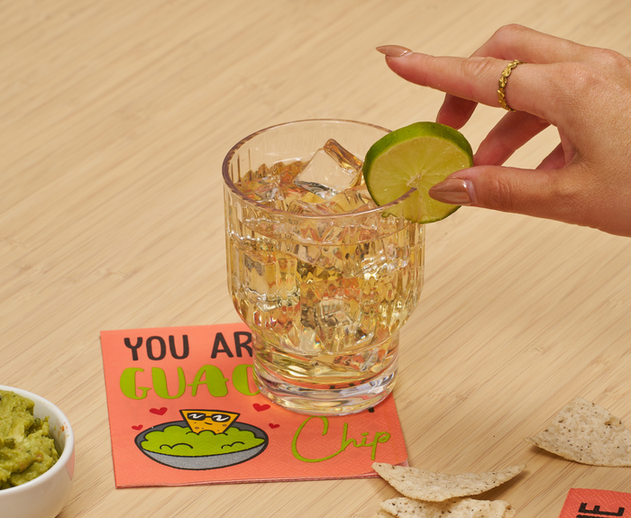 A hand with painted nails and a gold ring places a lime on a glass of clear drink atop Sugarplum’s "You are the GUAC to my Chip" Cocktail Napkins (26pk), beside tortilla chips and guacamole.