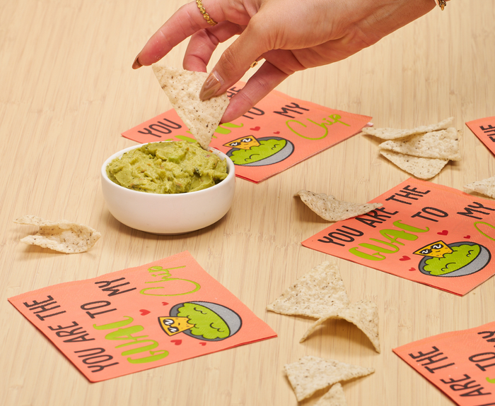 A hand dips a tortilla chip into guacamole. Sugarplum’s “You are the GUAC to my Chip” Cocktail Napkins (26pk) and several chips are scattered on a wooden surface, brightening the scene with their playful avocado design.
