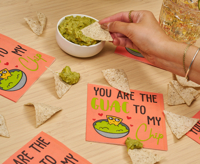 A hand dips a tortilla chip into guacamole beside Sugarplum’s “You are the GUAC to my Chip” cocktail napkins (26pk), scattered chips, and a glass of water, creating a festive, inviting scene.