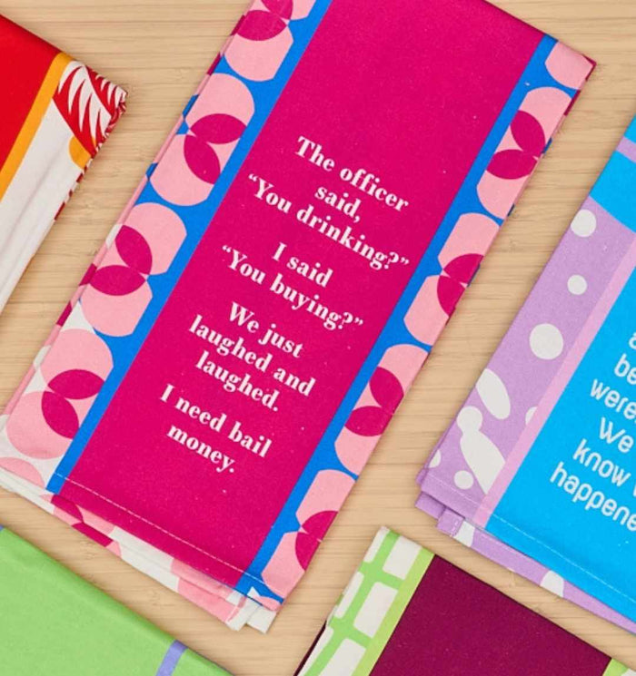 The "You Drinking? Dish Towel" by Drinks On Me features a funny quote perfect for the kitchen: "The officer said, You drinking? I said, You buying? We just laughed and laughed. I need bail money." Great as a humorous gift; other patterned towels are nearby.
