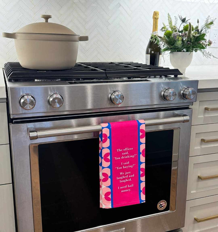 A modern stainless steel oven with a Drinks On Me “You Drinking?” dish towel hangs on the door, a beige Dutch oven sits on the stovetop, with a champagne bottle and floral arrangement on the white countertop in a bright kitchen.