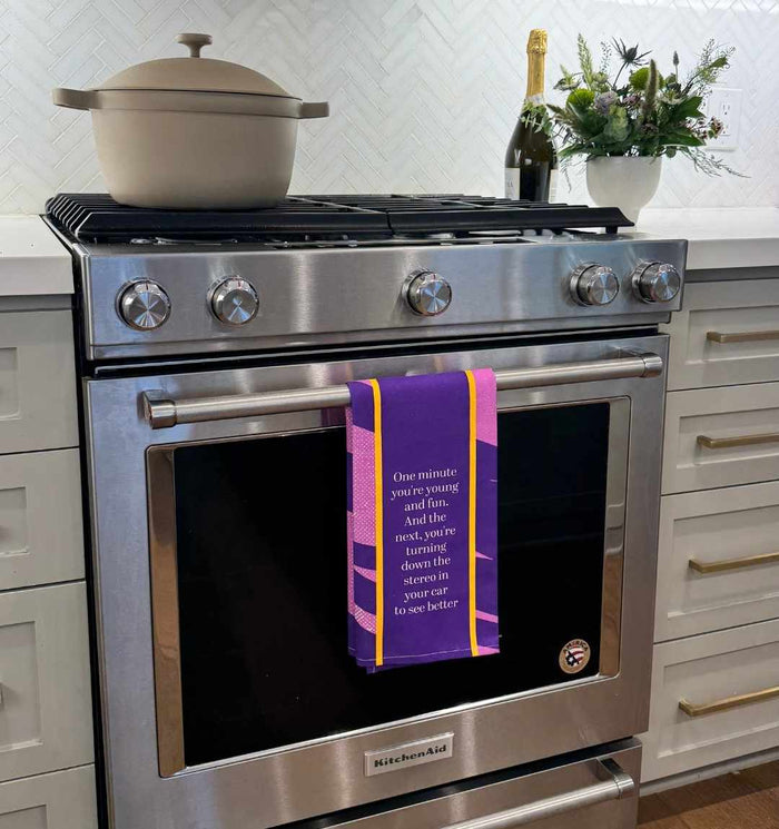 A modern stainless steel oven with the Drinks On Me Young and Fun Dish Towel, displaying a humorous quote, hangs on the handle. A beige pot sits on the stove, while a plant and champagne bottle rest on the countertop nearby.