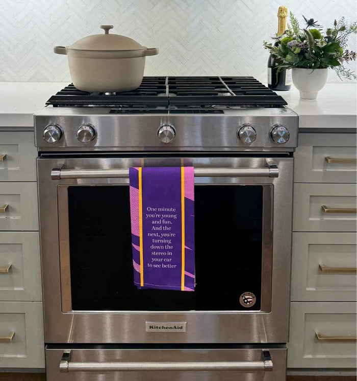 A modern stainless steel KitchenAid oven with a beige pot on top. The Drinks On Me "Young and Fun" dish towel hangs from the oven door. A bottle and a vase of flowers sit on the nearby white countertop.