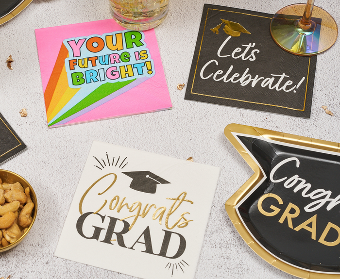 A flat lay of Sugarplum’s Congrats Grad Cocktail Napkins (26pk) with celebratory messages, plates reading “Congrats GRAD,” gold-rimmed glasses, and a bowl of nuts arranged on a white surface.