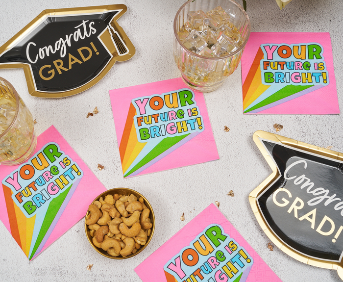 Colorful graduation party setup featuring Sugarplum's "Your future is bright" Cocktail Napkins (26pk), black and gold Congrats Grad! plates, drinks in glasses, cashews in a bowl, and confetti on a white surface.