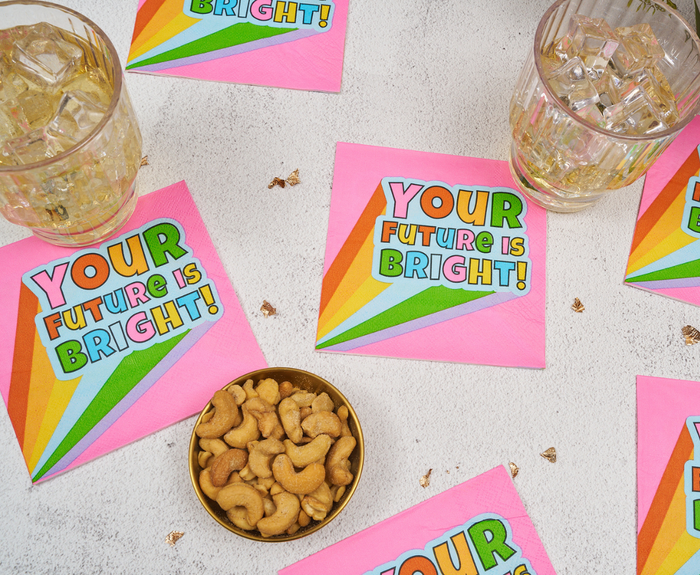 Sugarplum’s “Your future is bright” Cocktail Napkins (26pk) in pink with a rainbow design are shown beside two iced drinks and a bowl of cashews on a light surface.