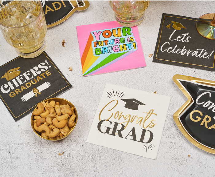A festive graduation party setup featuring Sugarplum Congrats Grad Cocktail Napkins (26pk), a bowl of cashews, plastic cups, and plates. The napkins are decorated with celebratory graduation messages.